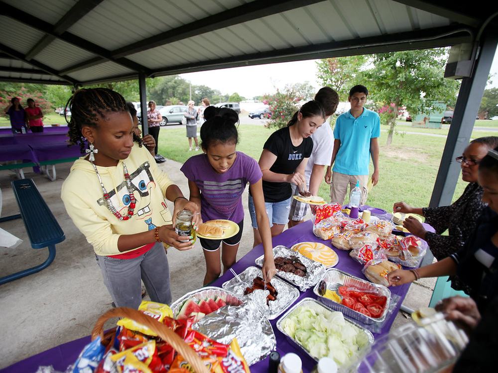 Young people help themselves at a long table of shared food outdoors.