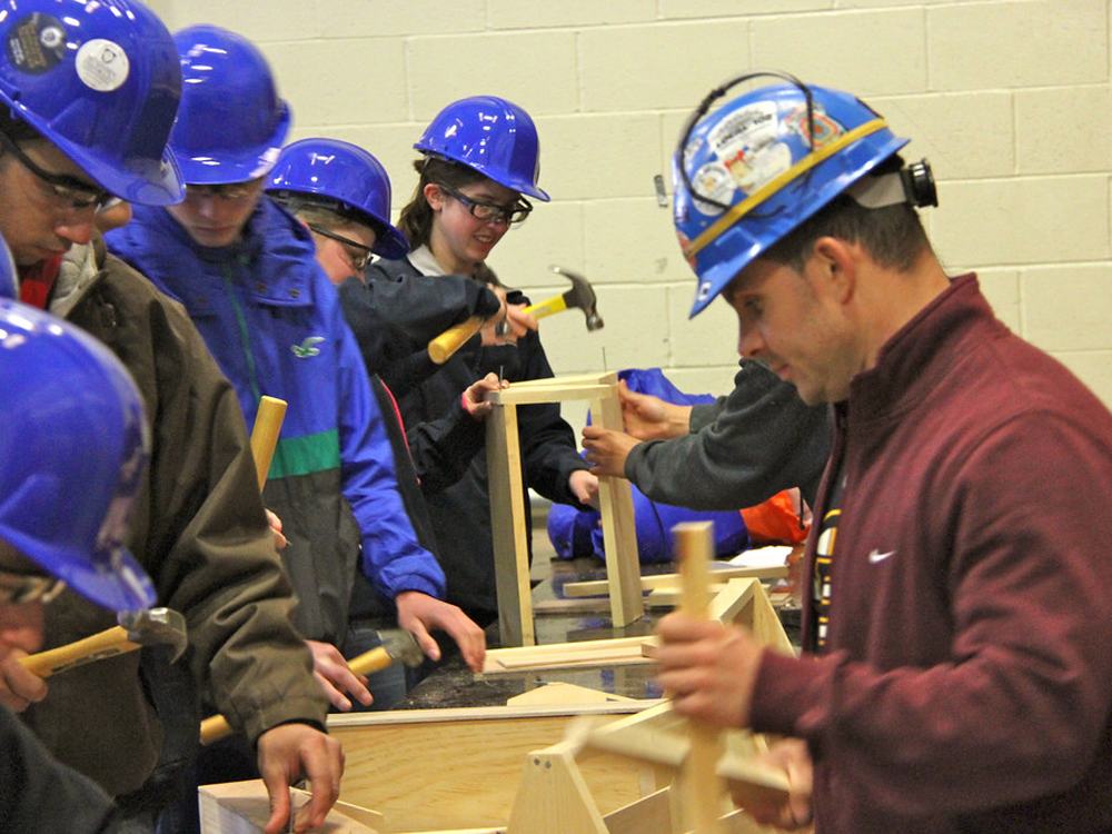 Teenagers in hard hats assemble a wooden frame beside an instructor.